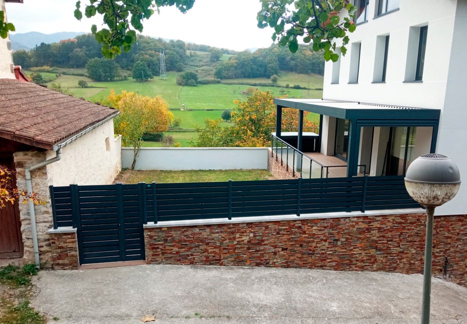 Cerramiento de lamas con puerta peatonal y barandillas de acero y vidrio.